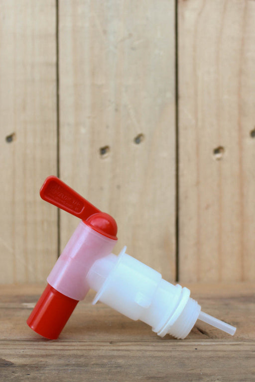 A plastic dispenser or faucet component with a red handle and red end, a translucent pink middle section, a white threaded body, and a clear tube, photographed on a wooden surface against a wood plank background.