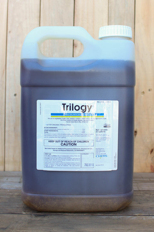 A large plastic jug of Trilogy with a white cap, sitting on a wooden surface against a light wood plank background.