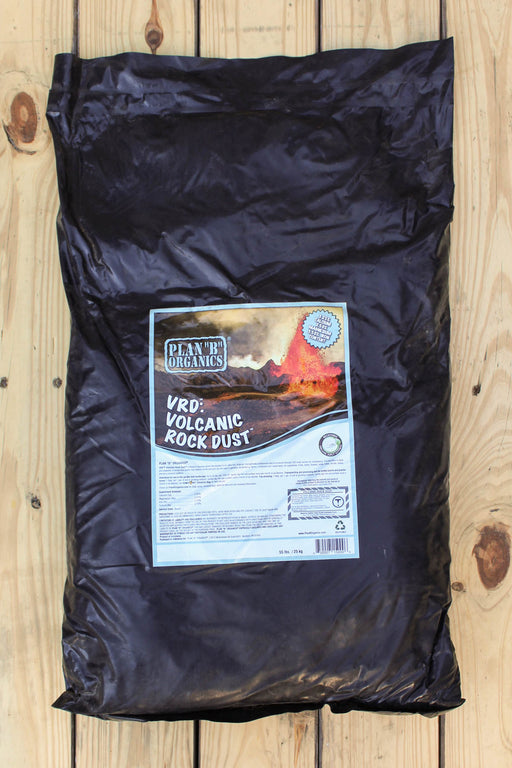 A large black plastic bag of VRD Volcanic Rock Dust, featuring a white and blue label with an illustration of a volcanic landscape, laid flat on a light-colored wooden plank surface.