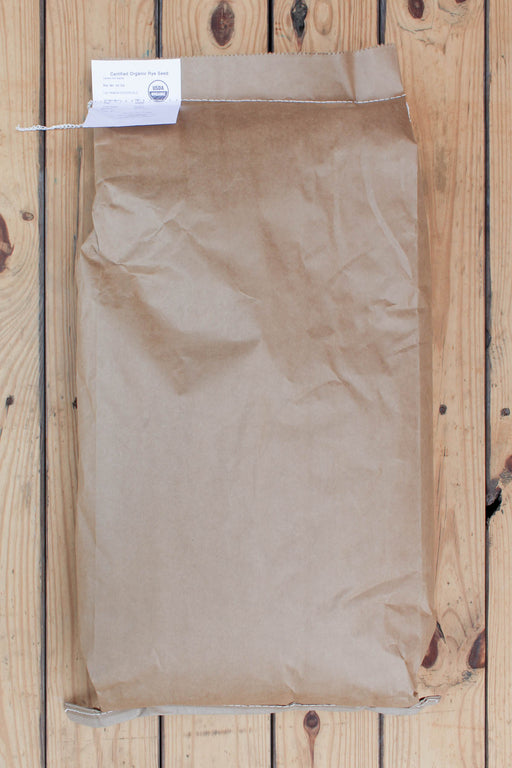 A brown kraft paper bag of winter rye with a white paper tag attached at the top, laid flat on a light wood plank surface.