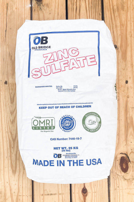 A white paper bag of Old Bridge Chemicals Zinc Sulfate, 25 kg, laid flat on a light-colored wooden plank surface. 
