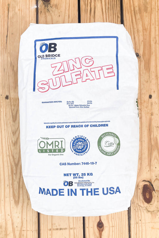 A white paper bag of Old Bridge Chemicals Zinc Sulfate, 25 kg, laid flat on a light-colored wooden plank surface. 